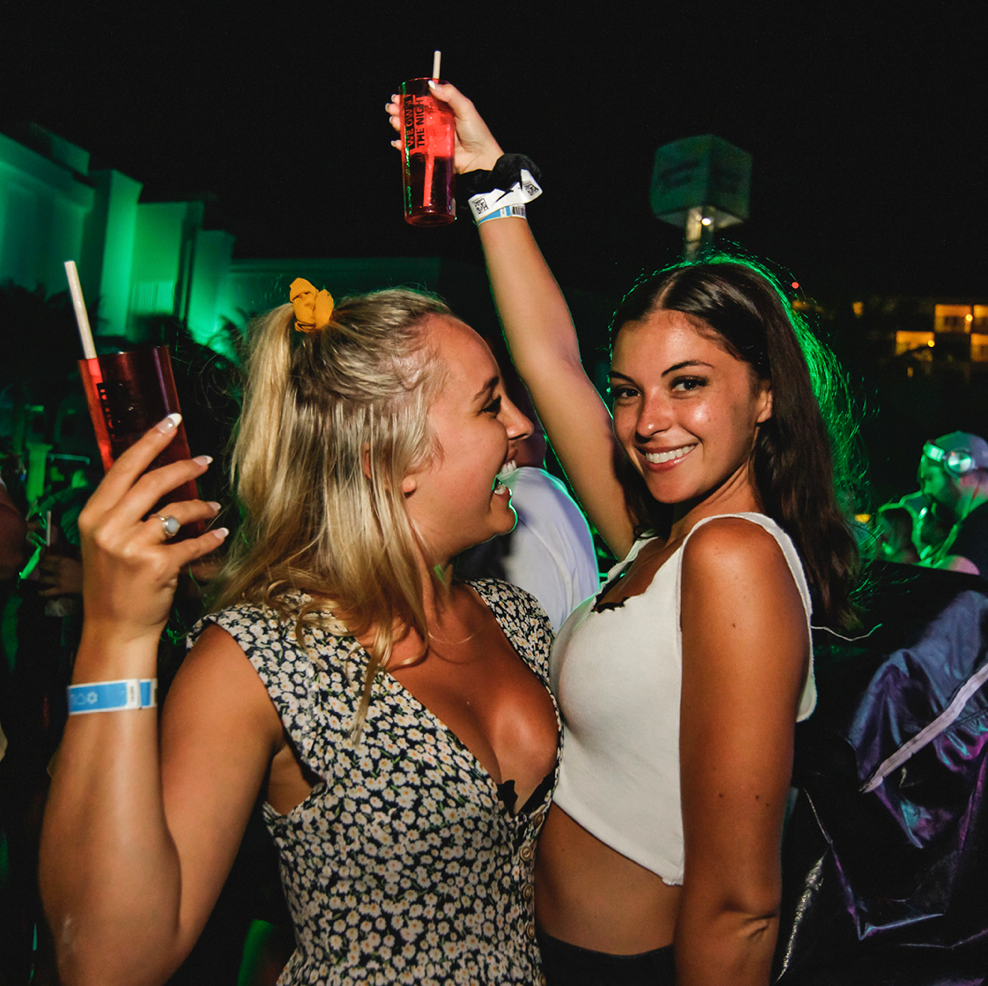 Two friends dancing and holding drinks under green neon lights at La Vaquita Cancún