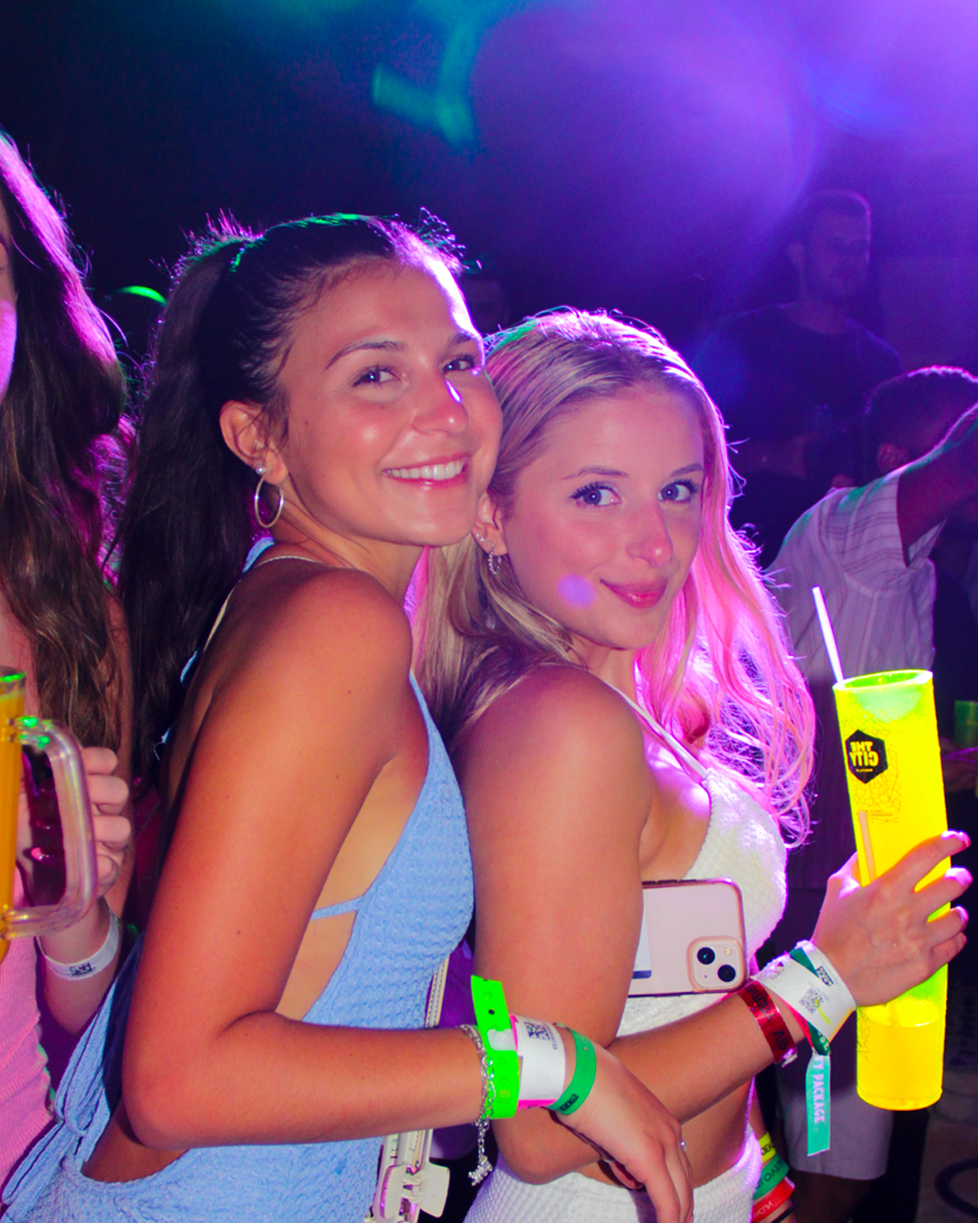 Two friends smiling and holding drinks under neon lights at Madness Tour Cancún