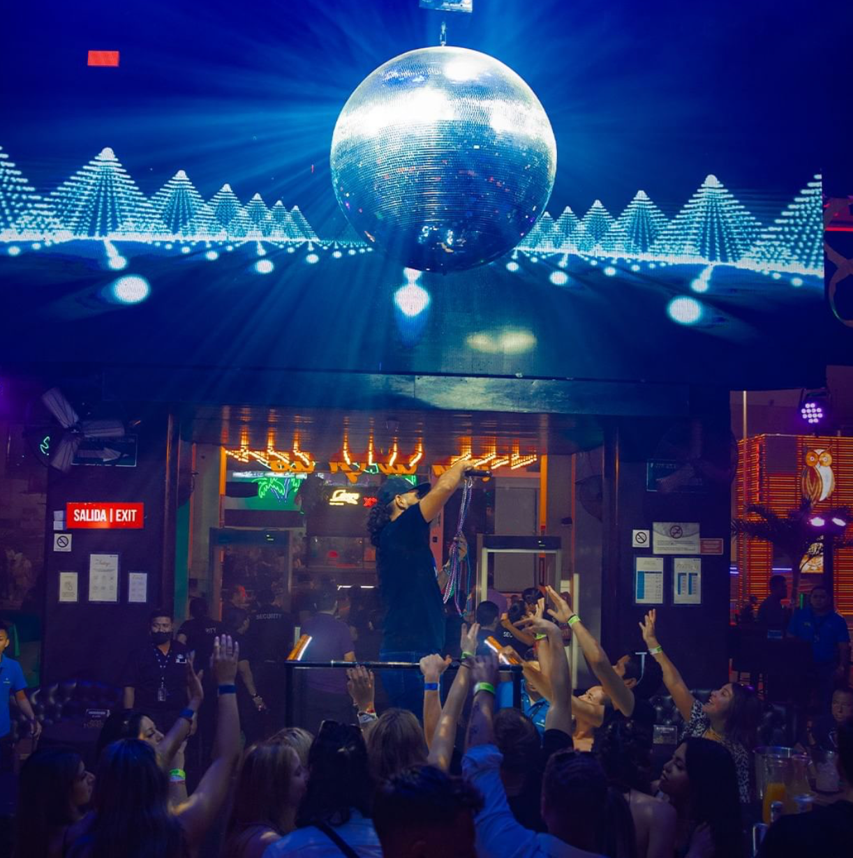 DJ performing under a giant disco ball with crowd raising hands at Mandala Club Cancún