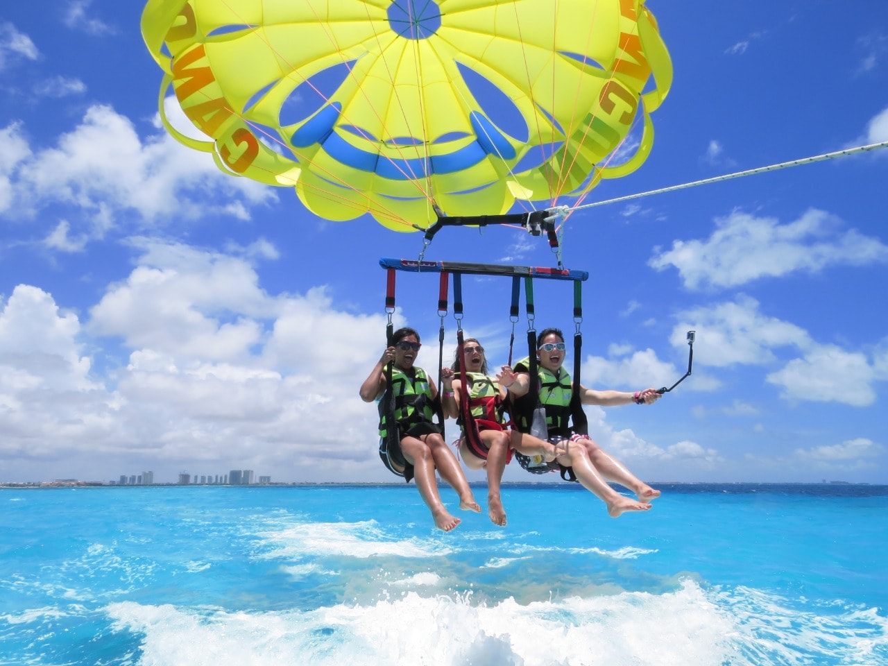 Three friends laughing and screaming with joy while parasailing over turquoise Cancun waters