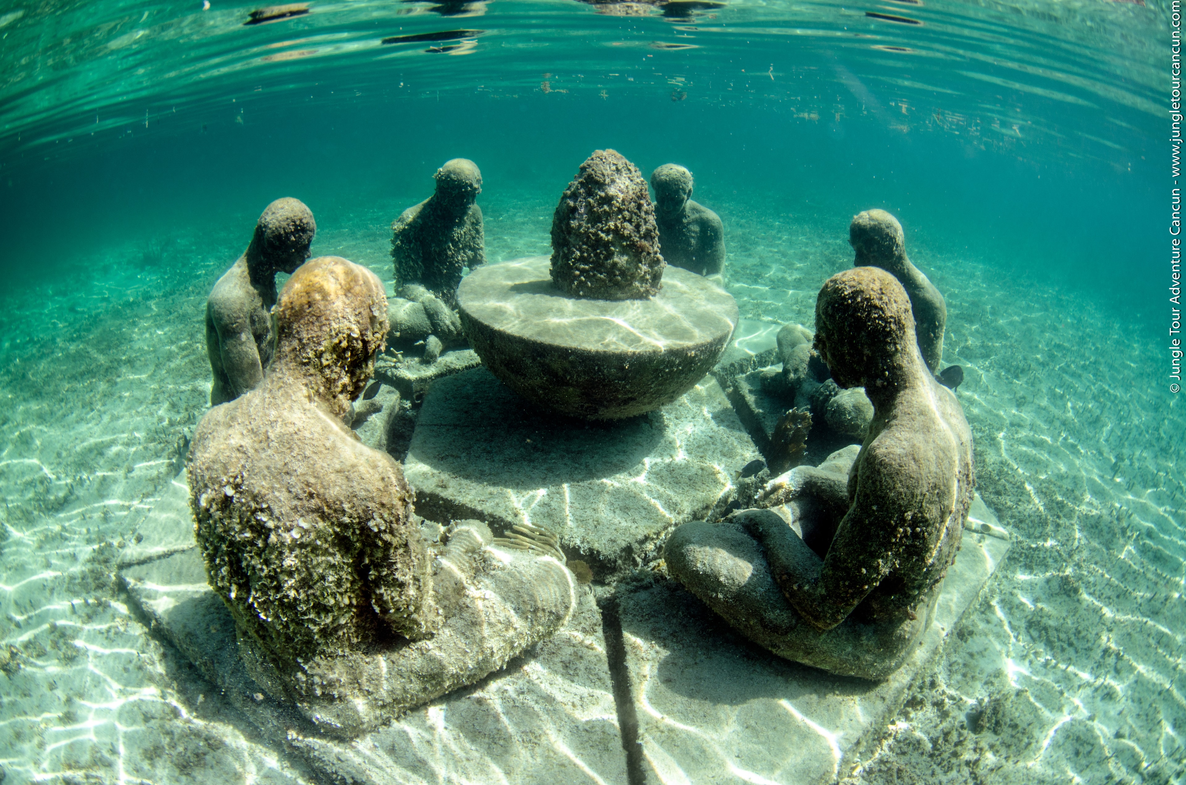 Underwater sculpture garden with coral-encrusted human figures seated around a table on the sandy seafloor in Cancun