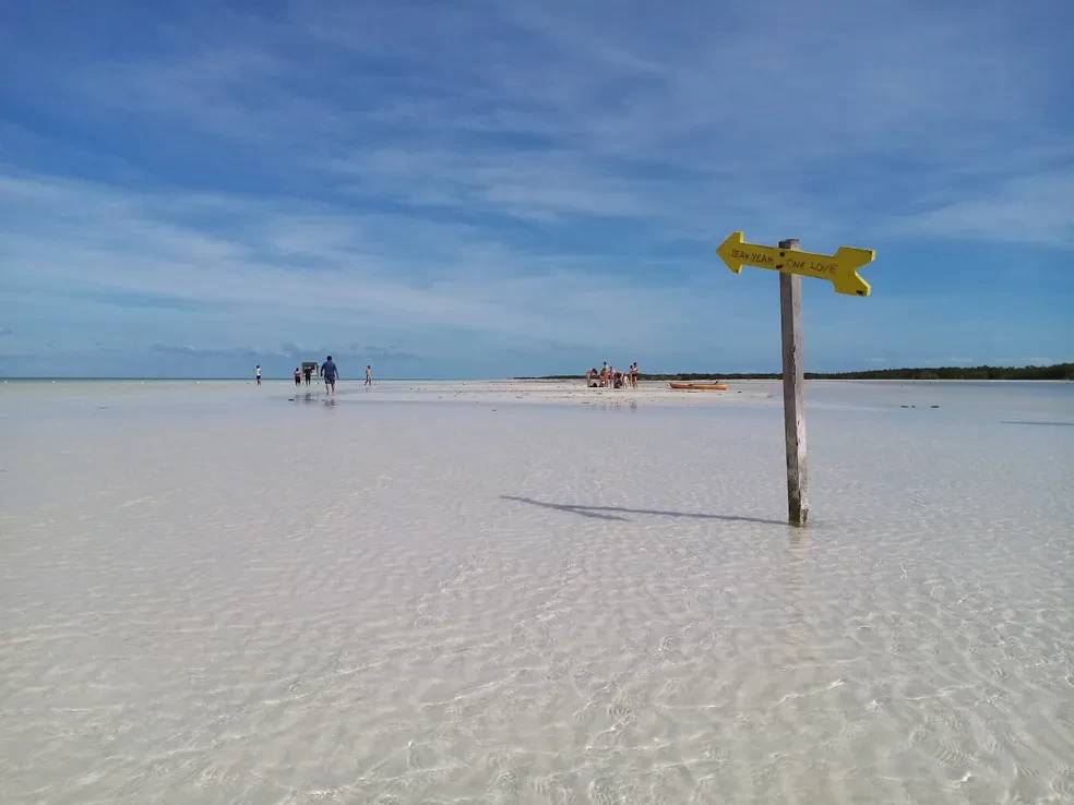 Shallow crystal-clear waters of Holbox with a yellow "Yeah Yeah One Love" directional sign and visitors wading in the distance