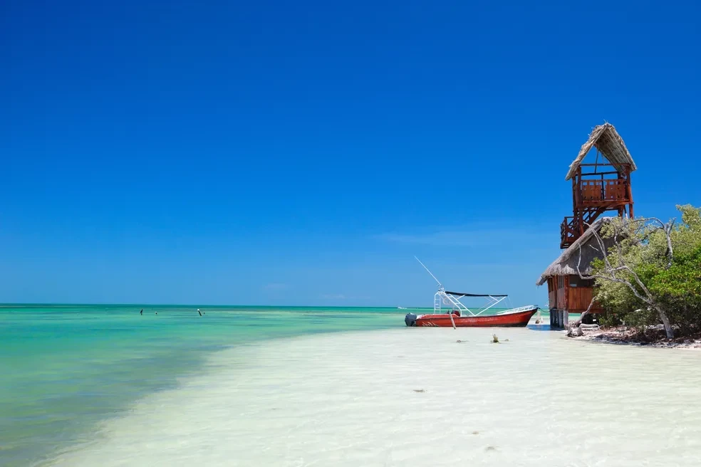 Red fishing boat moored beside a rustic thatched-roof tower on the white-sand shore of Holbox Island