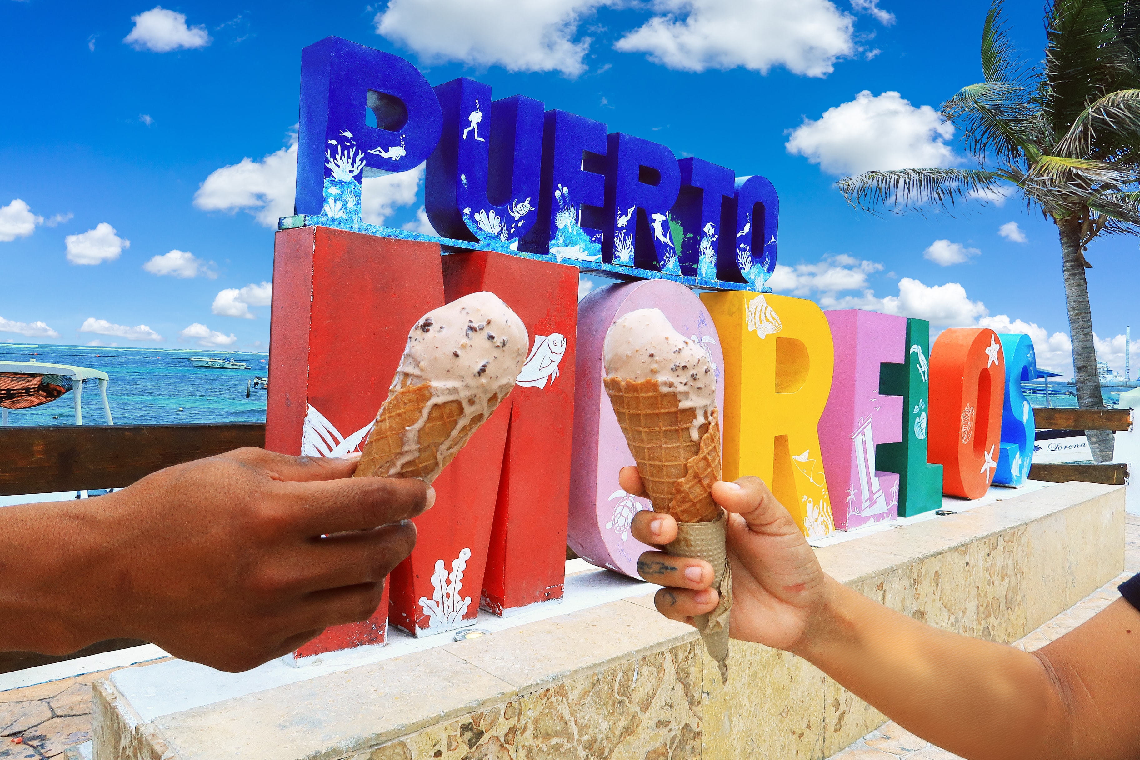 Two ice cream cones held up in front of the colourful PUERTO MORELOS letter sign on the beach with turquoise water behind