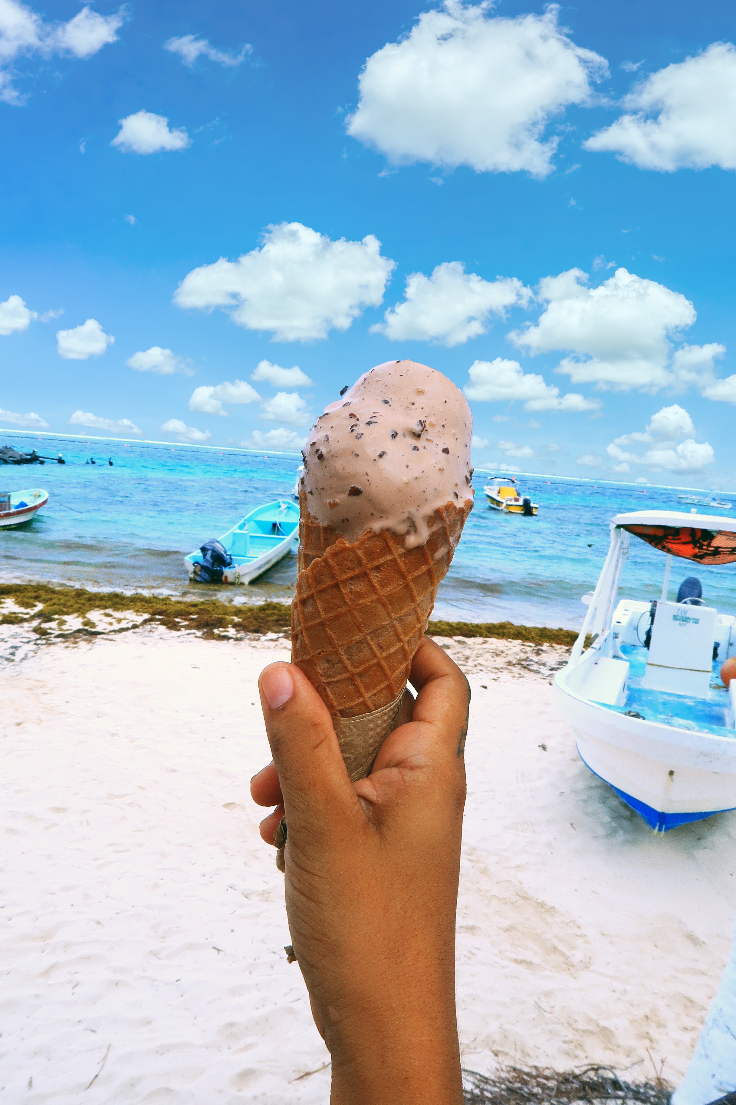 Ice cream cone held up against the white-sand beach and turquoise Caribbean water of Puerto Morelos with fishing boats behind