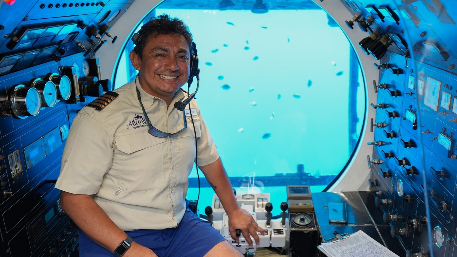 Smiling Atlantis Submarines Cozumel captain at the controls with tropical fish visible through the porthole behind him