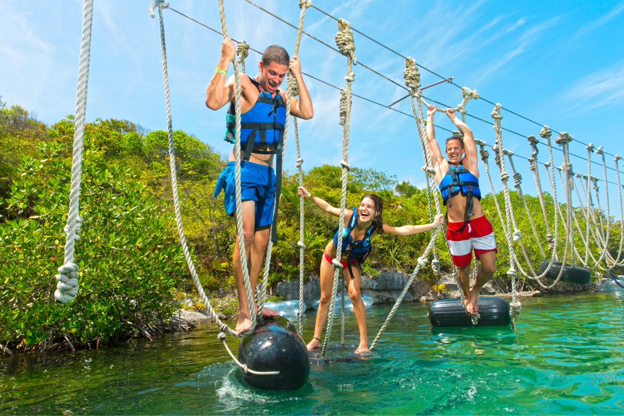 Three friends laughing on a rope obstacle course over turquoise water at Xel-Há
