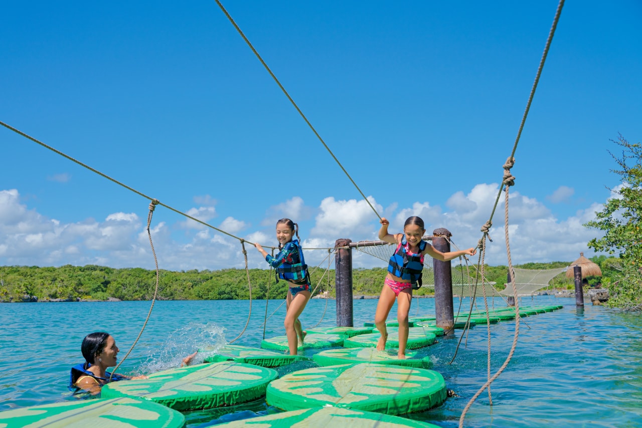 Two girls crossing a floating rope bridge over turquoise water at Xel-Há with their mother watching from below