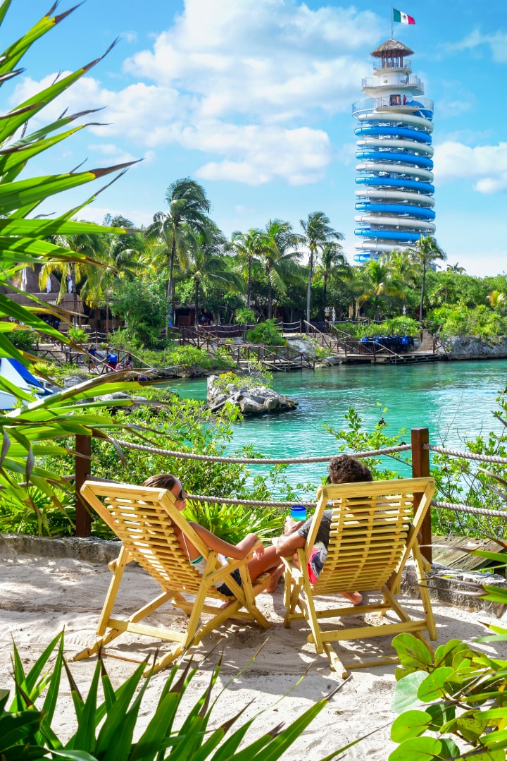 Couple relaxing on wooden sun loungers overlooking the Xel-Há inlet and spiral lighthouse tower