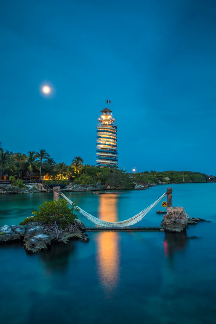 Xel-Há lighthouse tower illuminated at dusk with a hammock over the water and full moon above
