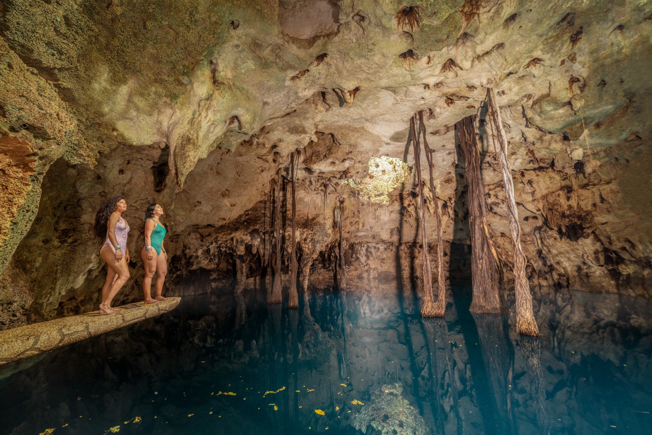 Two women standing on a ledge inside a dramatic cave cenote with turquoise water and hanging tree roots at Xenotes Oasis Maya