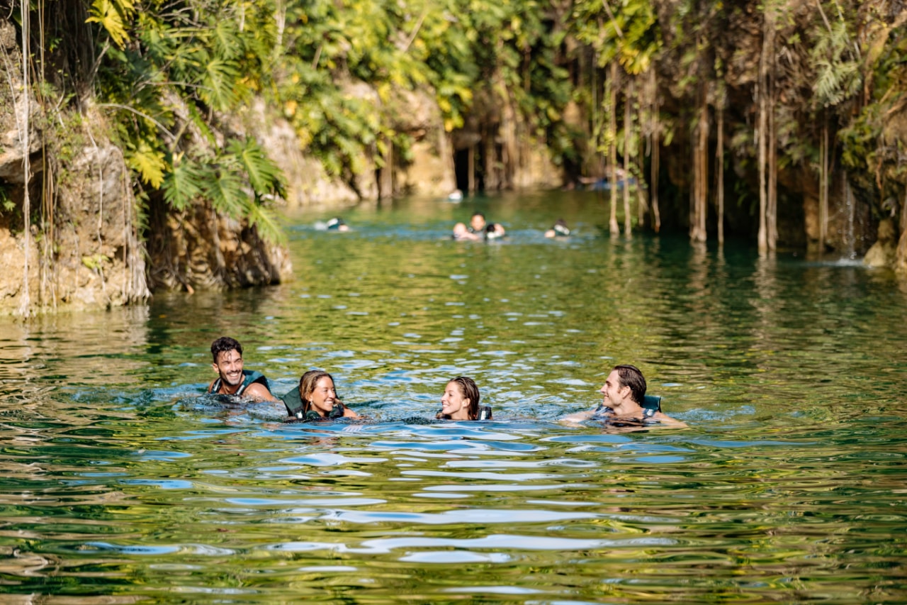Four friends swimming and laughing together in a lush open cenote channel at Xenotes Oasis Maya