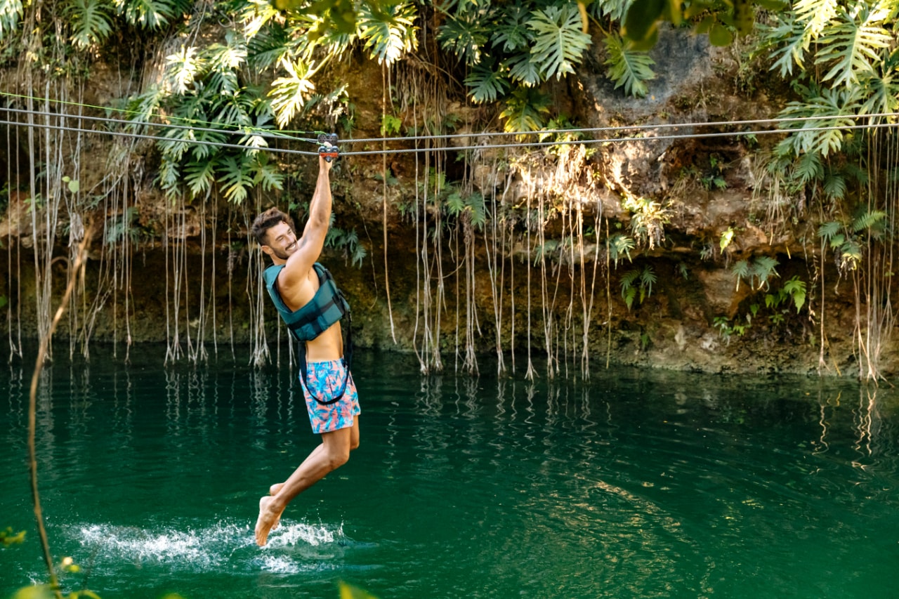 Man zip-lining over a turquoise cenote surrounded by hanging vines and tropical jungle at Xenotes Oasis Maya