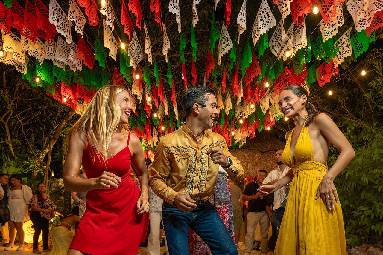Three friends dancing under red, white and green papel picado banners with string lights at Xoximilco