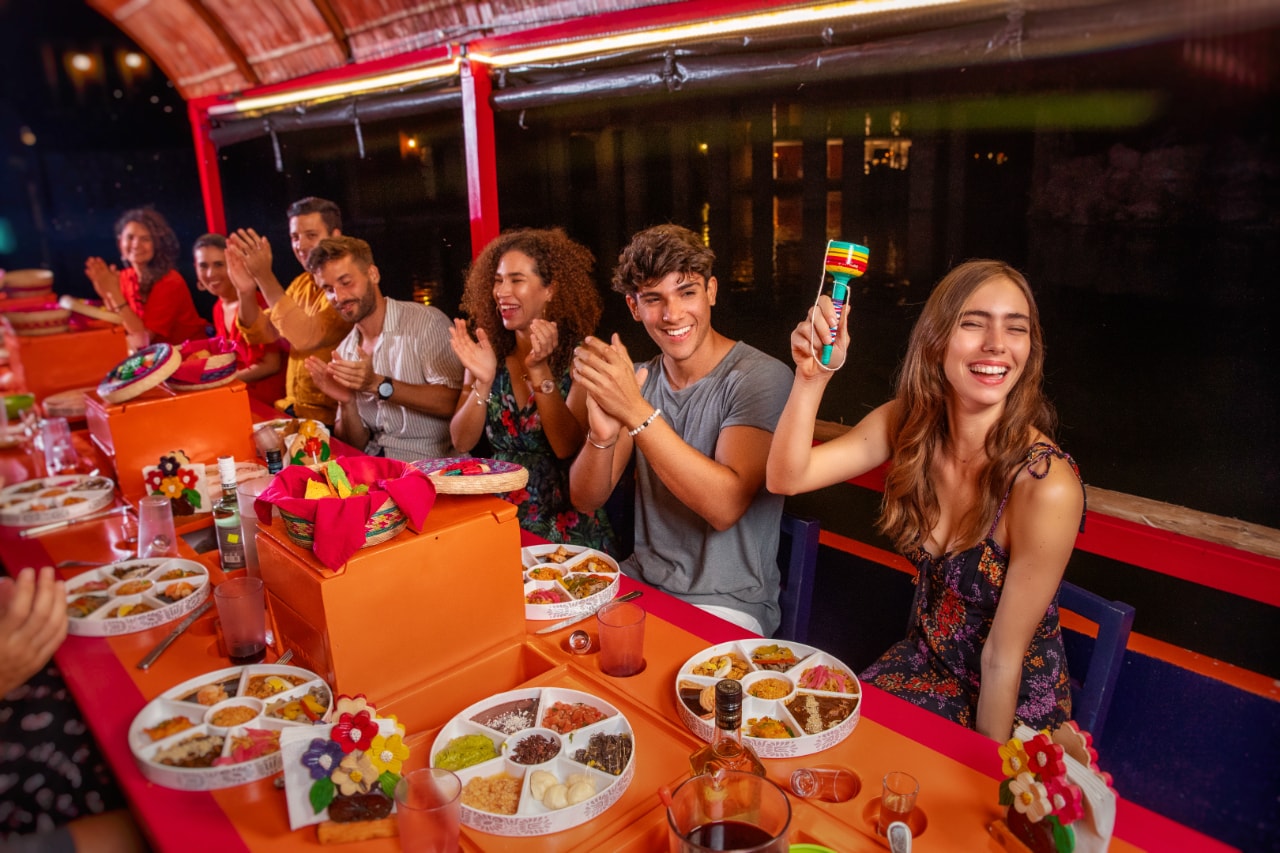 Guests clapping and celebrating at a festive dinner table on the Xoximilco boat with Mexican food and maracas